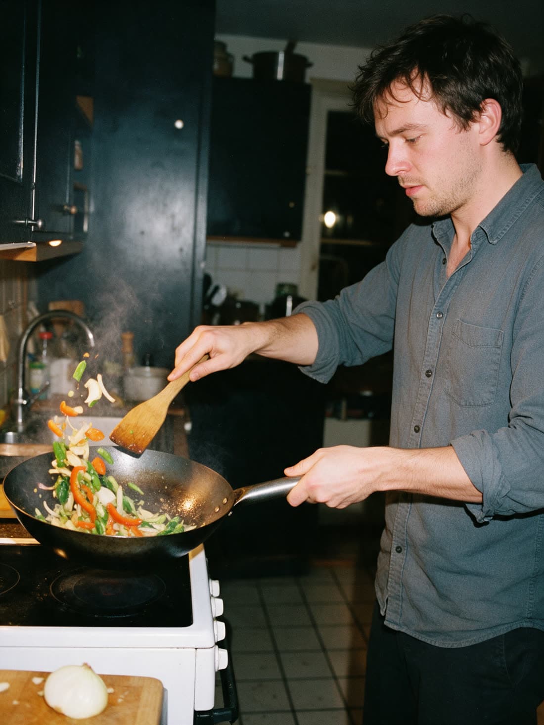 A man tossing vegetables in a wok at a home stove at night, spatula in one hand, the wok handle in the other